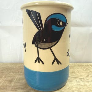 May include: A ceramic utensil holder with a hand-painted bird design. The cylindrical holder features a cream-coloured upper section, a blue lower section, and a black and blue bird illustration. Small, stylized footprints are scattered around the bird.