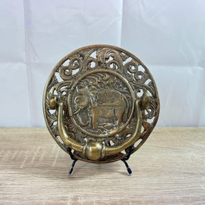 May include: Antique brass door knocker with an elephant design. The round knocker features an ornate, openwork border and a central relief of an elephant. A curved handle is attached below the elephant image.