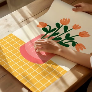 May include: A hand holding a pink and white illustration of a potted plant with orange flowers on a yellow grid background.