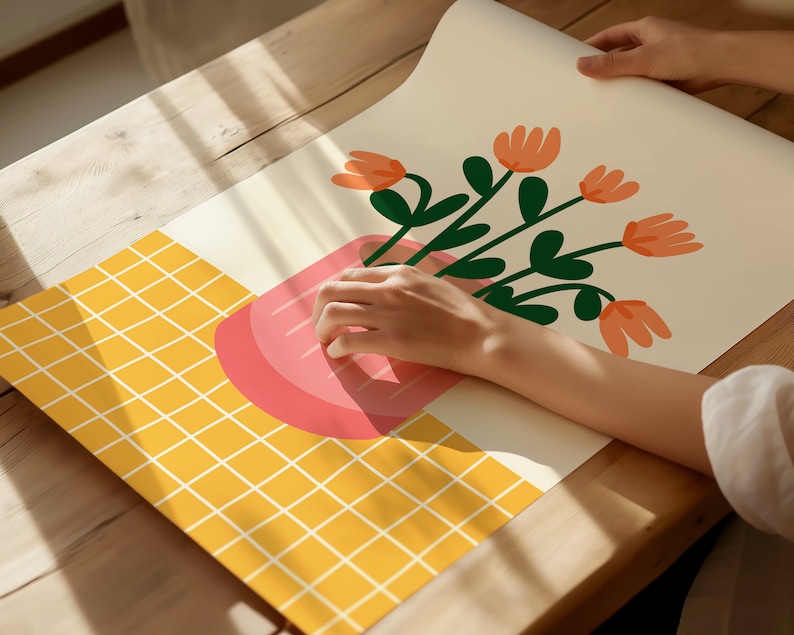 May include: A hand holding a pink and white illustration of a potted plant with orange flowers on a yellow grid background.