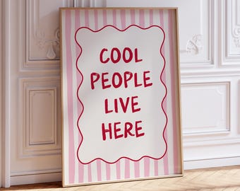 May include: A framed poster with the text "COOL PEOPLE LIVE HERE" in red, set against a white background with a wavy red border. The background has pink and white vertical stripes. The frame is a light brown color.