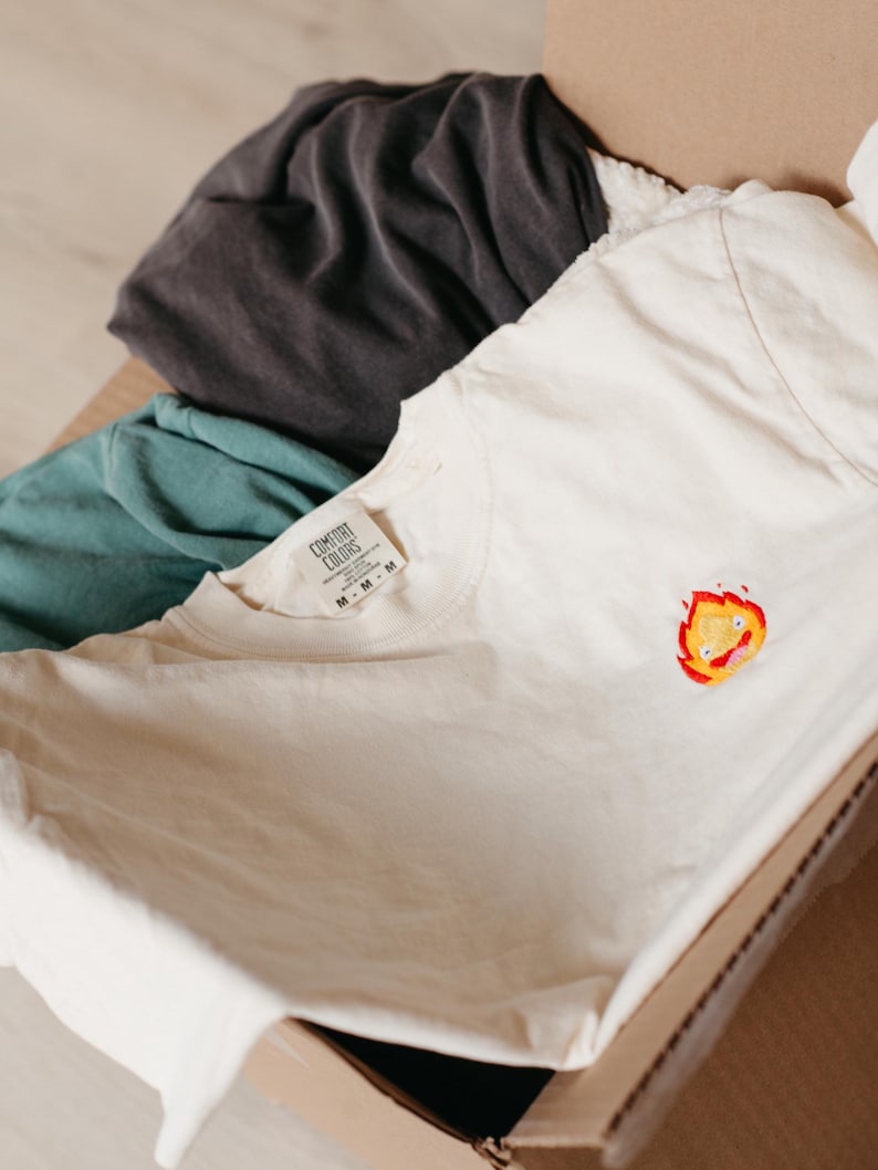 Puede incluir: Una camiseta blanca con una peque&ntilde;a imagen bordada de una llama amarilla y roja con una cara. La camiseta tiene una etiqueta que dice "COMFORT COLORS" y "M".