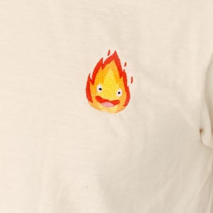 Puede incluir: Camiseta blanca con una imagen bordada de un esp&iacute;ritu de fuego amarillo y naranja con una cara sonriente.