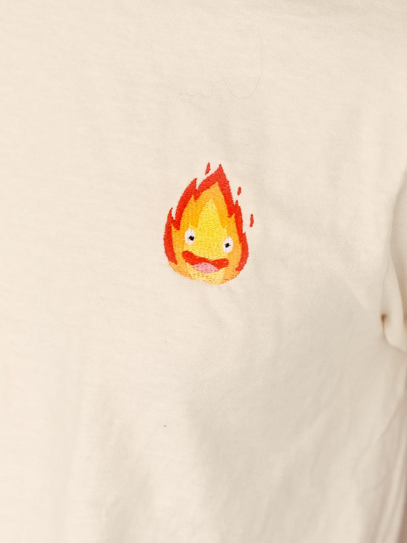 Puede incluir: Camiseta blanca con una imagen bordada de un esp&iacute;ritu de fuego amarillo y naranja con una cara sonriente.