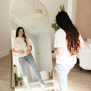 Puede incluir: Una persona de pie frente a un espejo arqueado de cuerpo entero, vistiendo una camiseta blanca con un peque&ntilde;o gr&aacute;fico naranja y pantalones vaqueros azules. El espejo est&aacute; de pie sobre un suelo de madera.