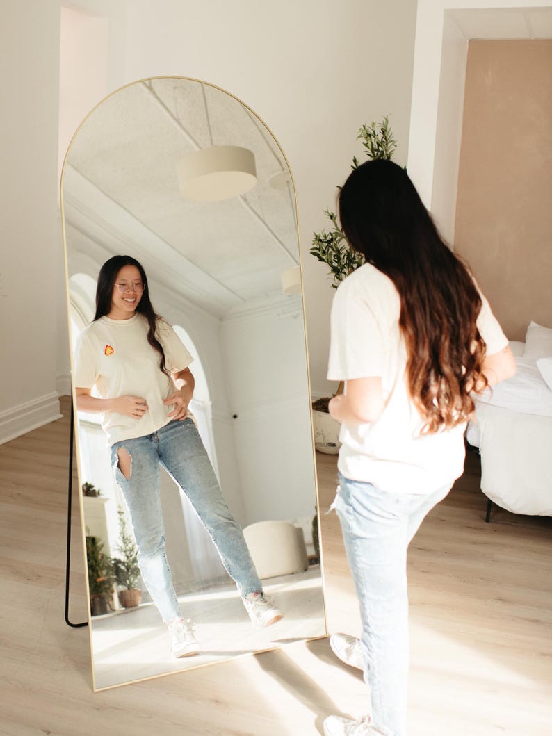 Puede incluir: Una persona de pie frente a un espejo arqueado de cuerpo entero, vistiendo una camiseta blanca con un peque&ntilde;o gr&aacute;fico naranja y pantalones vaqueros azules. El espejo est&aacute; de pie sobre un suelo de madera.