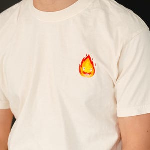 May include: A white t-shirt with a small embroidered image of a yellow fire spirit with a red flame. The spirit has a smiling face and is wearing a red tongue.