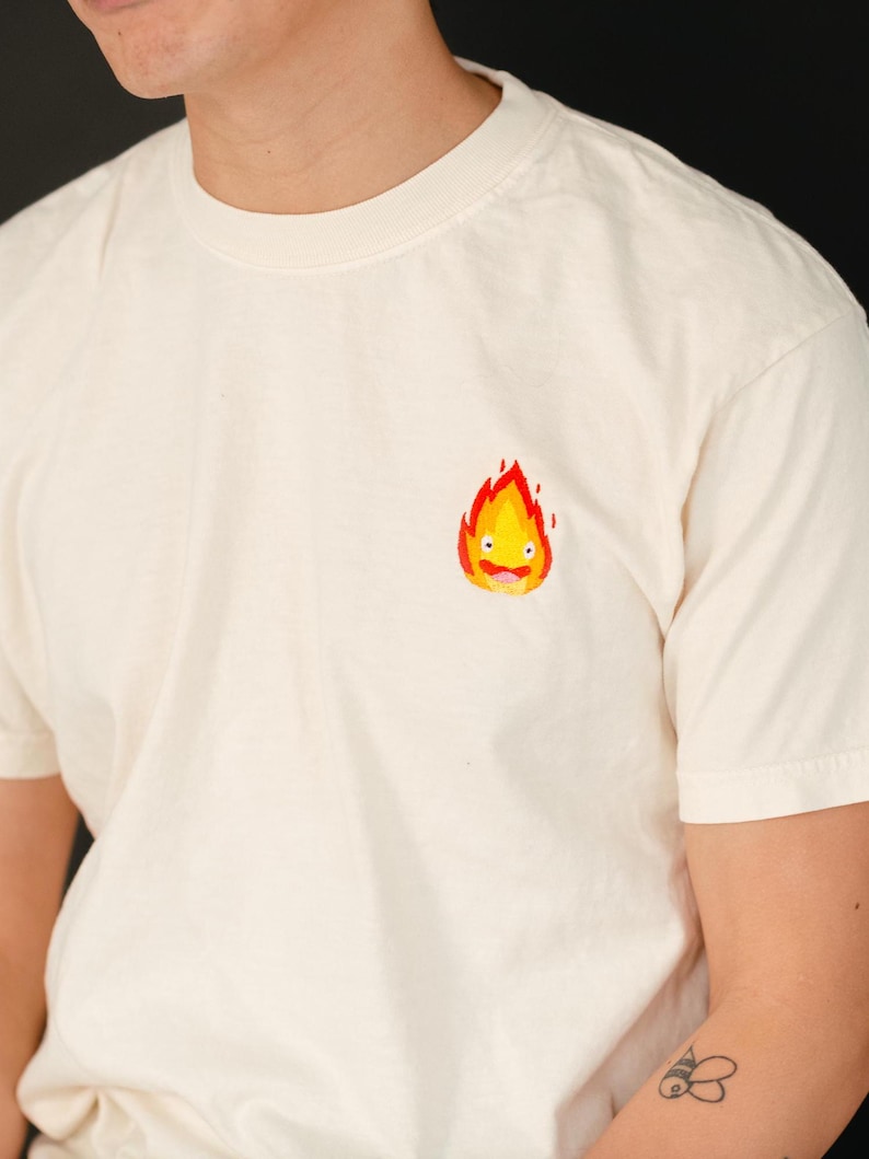 Puede incluir: Una camiseta blanca con una peque&ntilde;a imagen bordada de un esp&iacute;ritu de fuego amarillo con una llama roja. El esp&iacute;ritu tiene una cara sonriente y lleva una lengua roja.
