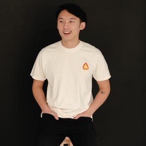 Puede incluir: Una persona con una camiseta blanca con un peque&ntilde;o gr&aacute;fico de llama naranja en la parte delantera. La persona est&aacute; sentada en un taburete de madera y tiene un peque&ntilde;o tatuaje en el brazo.