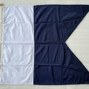 Op de afbeelding: Een nautische signaalvlag met een verticaal verdeeld ontwerp, bestaande uit een wit linkergedeelte en een donkerblauw rechtergedeelte dat eindigt in een zwaluwstaartpunt. De vlag is voorzien van een beige canvas broeking met een houten knevel.