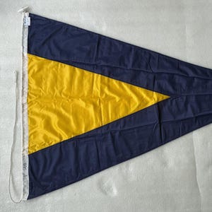 Op de afbeelding: Een driehoekige nautische signaalvlag met een grote gele driehoek in het midden op een donkerblauwe achtergrond. De vlag heeft een witte verticale zijde met een touwbevestiging en de tekst 1ST SUB in zwarte inkt op de witte stof.