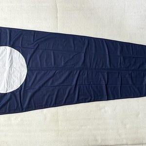 Op de afbeelding: Een lange, driehoekige marineblauwe wimpel met een grote, effen witte cirkel nabij het bredere uiteinde. De vlag heeft een witte verticale kopstrip aan de linkerzijde met twee kleine ogen voor bevestiging en een puntig uiteinde.