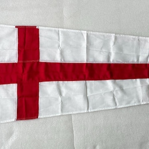 Op de afbeelding: Een witte driehoekige wimpel met een opvallende rode horizontale baan door het midden en een verticale rode baan bij de mastzijde, die een kruispatroon vormt. De vlag is voorzien van een wit touwlusje aan de linkerzijde.