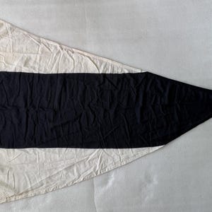 Op de afbeelding: Een driehoekige nautische signaalvlag met een brede horizontale zwarte baan gecentreerd tussen twee gebroken witte secties. De vlag is voorzien van een wit touw met een metalen clip aan de verticale hijskant voor een veilige bevestiging.