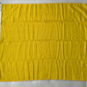 Op de afbeelding: Een effen gele rechthoekige vlag met een witte verticale kopband aan de linkerzijde. De vlag is voorzien van een wit touw aan de boven- en onderkant van de kopband, met een klein wit label zichtbaar nabij de bovenhoek.