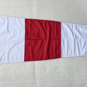 Op de afbeelding: Een driehoekige nautische wimpel met een herhalend patroon van afwisselende verticale rode en witte banen. De vlag is voorzien van een witte verstevigde broeking aan de linkerzijde met een witte touwlus voor bevestigingsdoeleinden.