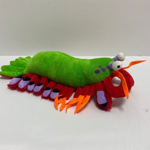 Puede incluir: Un juguete de peluche verde y rojo con forma de mantis. El juguete tiene ojos grandes y blancos y garras naranjas.