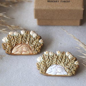 May include: Two ceramic brooches with a sleeping rabbit design, surrounded by leaves and flowers. The brooches are brown, white, and beige. A small, brown box with the text "BILBERRY FOREST" and a fox illustration is in the background.