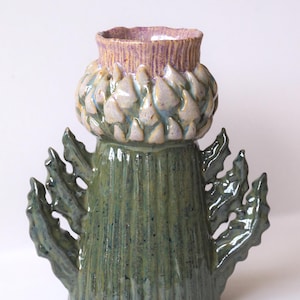 Pode incluir: Um vaso de cerâmica em forma de cardo com uma base verde texturizada e duas folhas laterais. A parte superior apresenta uma estrutura bulbosa e em camadas em tons de roxo claro e bege, com uma abertura estriada de cor lavanda.