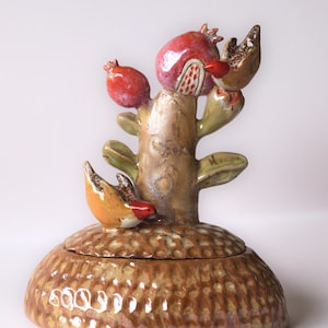 Puede incluir: Un tarro de cerámica con tapa y base abovedada de color tostado con textura. Un cactus esculpido se eleva desde el centro, adornado con granadas de color rojo vibrante y un pequeño pájaro naranja y rojo posado en el tallo del cactus.