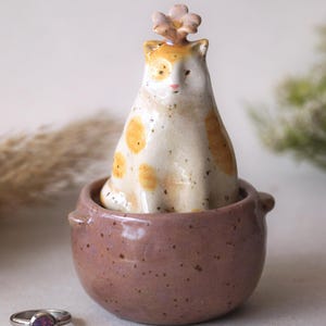Può includere: Una statuina di gatto in ceramica con macchie arancioni e un fiore sulla testa è posta all'interno di una piccola ciotola maculata color malva. Un anello d'argento con una pietra viola è appoggiato vicino. La statuina del gatto è un oggetto decorativo.