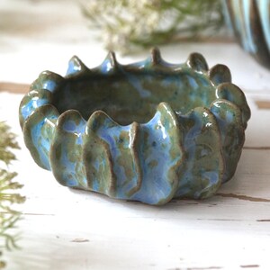 May include: A handmade ceramic bowl with a unique, wavy rim. The bowl features a blend of blue and green glaze, creating a textured, organic appearance. The bowl is likely used for decorative purposes or to hold small items.