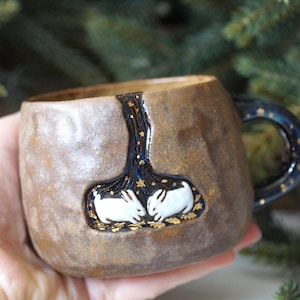 May include: A textured brown ceramic mug featuring a dark blue, star-filled inlay depicting two white rabbits facing each other. The handle is dark blue with gold star accents, and the overall finish of the mug has a rustic, earthy appearance.