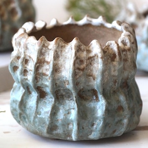 May include: A small, round ceramic planter with a textured, ribbed exterior. The surface features a mottled light blue and sage green glaze with exposed brown clay patches. The rim has a scalloped, uneven edge, and the interior is smooth.