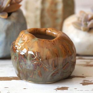 Pode incluir: Um pequeno vaso redondo de cerâmica marrom com uma superfície texturizada e uma abertura larga. O vaso tem um esmalte verde claro e marrom com um efeito manchado.