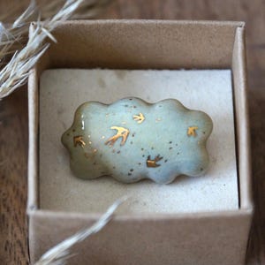 May include: A light blue cloud-shaped ceramic brooch with gold-colored bird accents, presented in a small cardboard box. The brooch has a glossy finish and is speckled with tiny dark dots. A unique, handcrafted accessory.