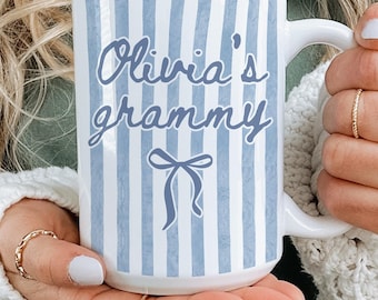Pode incluir: Caneca de cerâmica branca com listras verticais azuis e o texto "Olivia's grammy" em fonte cursiva. Um laço azul está abaixo do texto. A caneca tem uma alça branca.