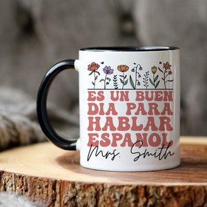 Może przedstawiać: Biały ceramiczny kubek do kawy z czarnym rantem i uchwytem. Kubek ma kwiatowy wz&oacute;r z tekstem "Es un buen dia para hablar espanol Mrs. Smith" w kolorze r&oacute;żowym.