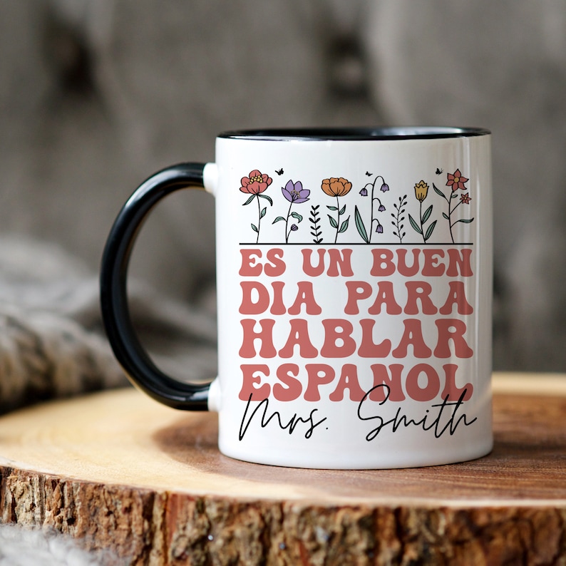 Może przedstawiać: Biały ceramiczny kubek do kawy z czarnym rantem i uchwytem. Kubek ma kwiatowy wz&oacute;r z tekstem "Es un buen dia para hablar espanol Mrs. Smith" w kolorze r&oacute;żowym.
