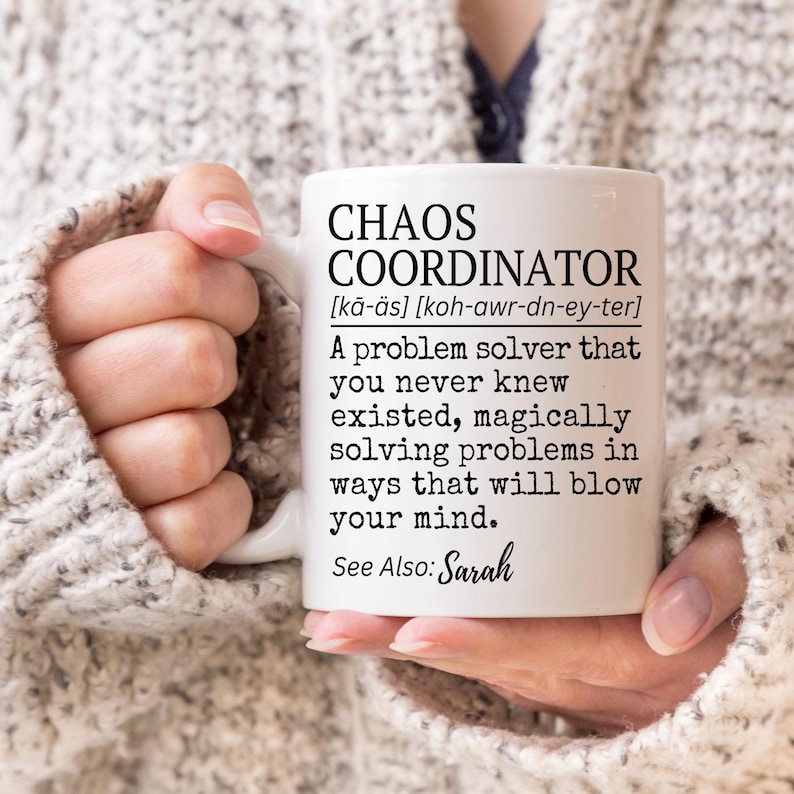 Puede incluir: Taza de cer&aacute;mica blanca con el texto "CHAOS COORDINATOR" y una definici&oacute;n. La taza tambi&eacute;n incluye el texto "See Also: Sarah". La taza est&aacute; sostenida por una persona que lleva un su&eacute;ter de color claro.