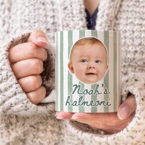 May include: White ceramic mug with a green and white striped pattern. The mug features a baby's face and the text "Noah's halmeoni" in green script. The mug is held by a person wearing a white sweater.