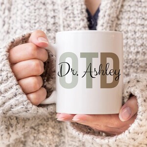 May include: White ceramic mug with the text "OTD Dr. Ashley" in black and brown lettering. The letters "OTD" are in a larger font size and are colored in shades of green and brown.