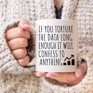 May include: White ceramic mug with the text "IF YOU TORTURE THE DATA LONG ENOUGH IT WILL CONFESS TO ANYTHING" in black. The mug also features a black bar graph with an upward arrow. The mug is held by a person wearing a gray sweater.