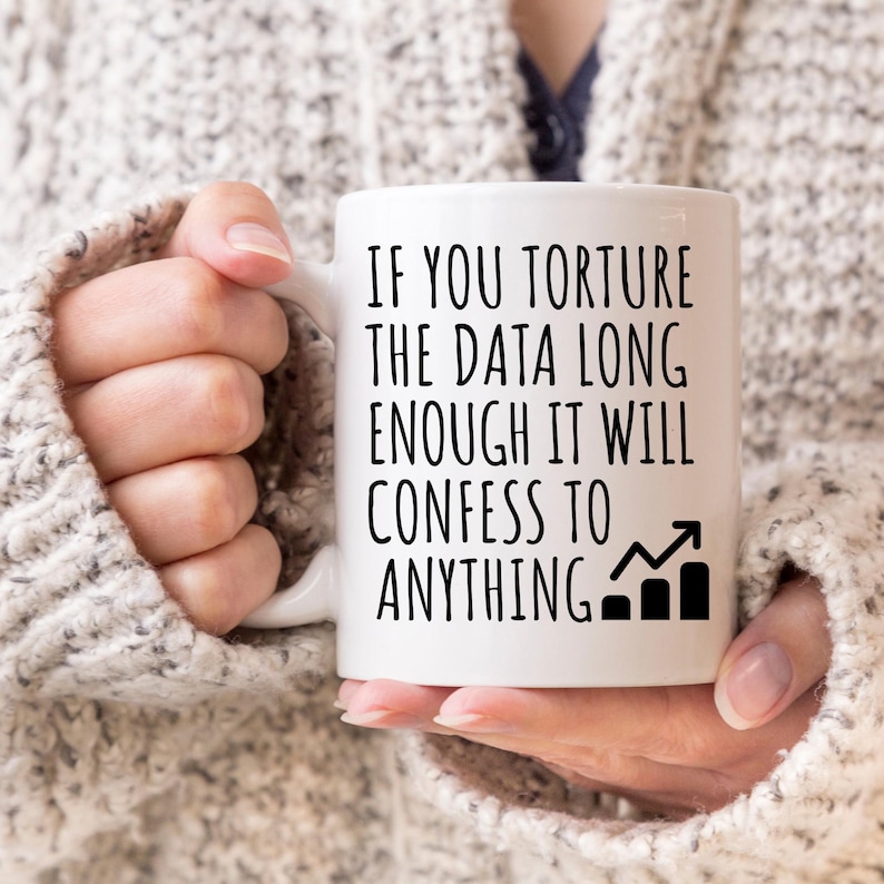 May include: White ceramic mug with the text "IF YOU TORTURE THE DATA LONG ENOUGH IT WILL CONFESS TO ANYTHING" in black. The mug also features a black bar graph with an upward arrow. The mug is held by a person wearing a gray sweater.