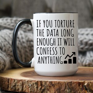 May include: White ceramic mug with a black handle and rim. The mug features the text "IF YOU TORTURE THE DATA LONG ENOUGH IT WILL CONFESS TO ANYTHING" in black, along with a graph icon.