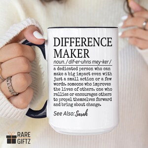 May include: White ceramic mug with a black interior and handle, featuring the text "DIFFERENCE MAKER" in black. The mug includes a definition of the term and the words "See Also: Sarah". The mug is held by a person.