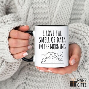 May include: White ceramic mug with a black handle and rim, featuring the text "I LOVE THE SMELL OF DATA IN THE MORNING" and a black and white line graph. The mug is held by a person wearing a grey sweater.