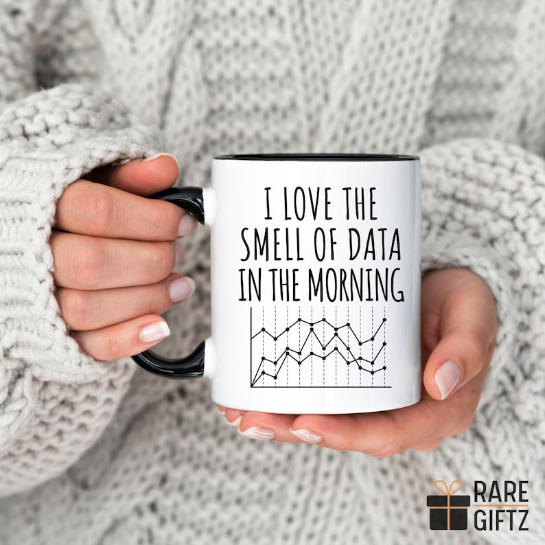 May include: White ceramic mug with a black handle and rim, featuring the text "I LOVE THE SMELL OF DATA IN THE MORNING" and a black and white line graph. The mug is held by a person wearing a grey sweater.