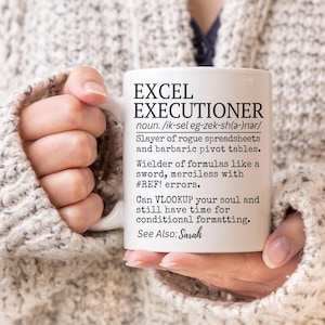 May include: White ceramic mug with the text "EXCEL EXECUTIONER" and a humorous definition. The mug is held by a person wearing a gray sweater. The text includes a pronunciation guide and a reference to Sarah.