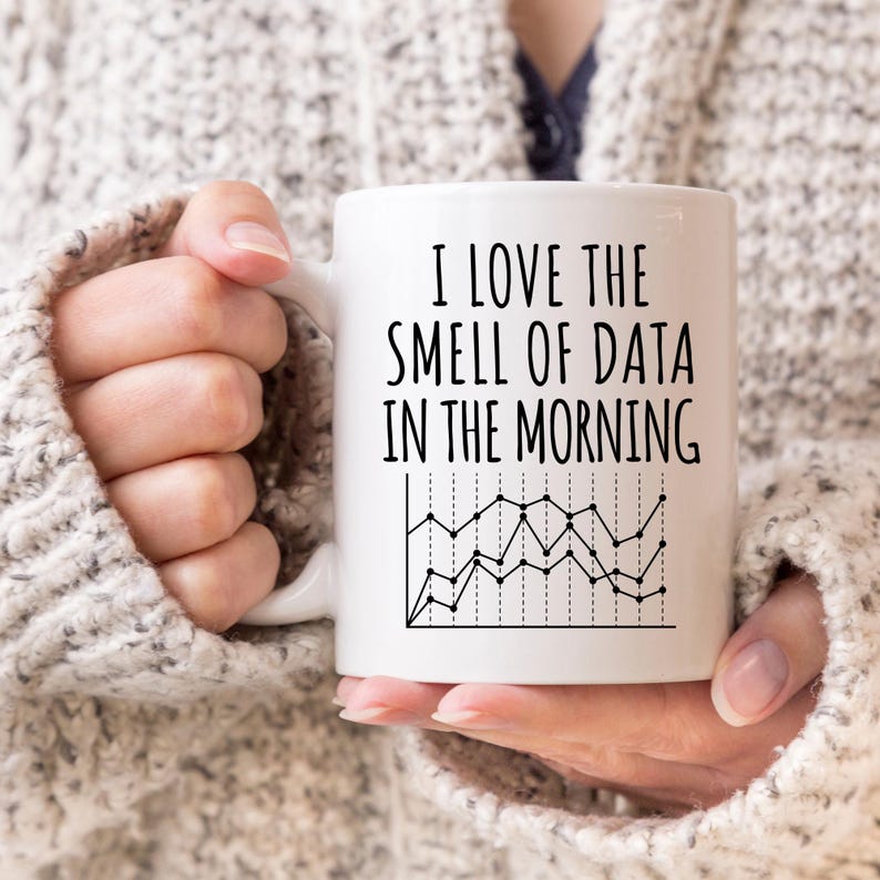 May include: White ceramic mug with the text "I LOVE THE SMELL OF DATA IN THE MORNING" in black, and a line graph illustration. The mug is held by a person wearing a grey sweater.