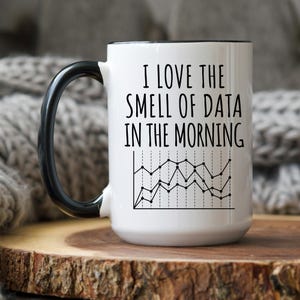 May include: White ceramic mug with a black handle and rim. The mug features the text "I LOVE THE SMELL OF DATA IN THE MORNING" above a black and white line graph design. The mug is on a wooden surface.