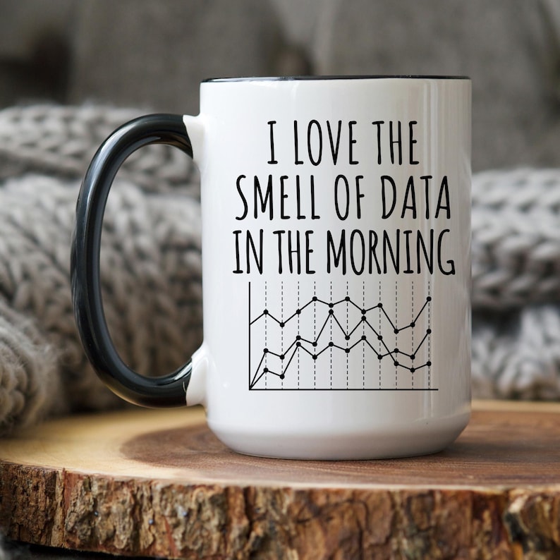 May include: White ceramic mug with a black handle and rim. The mug features the text "I LOVE THE SMELL OF DATA IN THE MORNING" above a black and white line graph design. The mug is on a wooden surface.