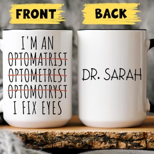 May include: White ceramic mug with a black handle and rim. The front of the mug has the text "I'M AN OPTOMATRIST OPTOMETREST OPTOMOTRYST I FIX EYES" with the words crossed out. The back of the mug says "DR. SARAH".
