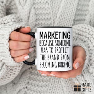 May include: White coffee mug with a black rim and handle. The mug has black text that reads "Marketing because someone has to protect the brand from becoming boring."