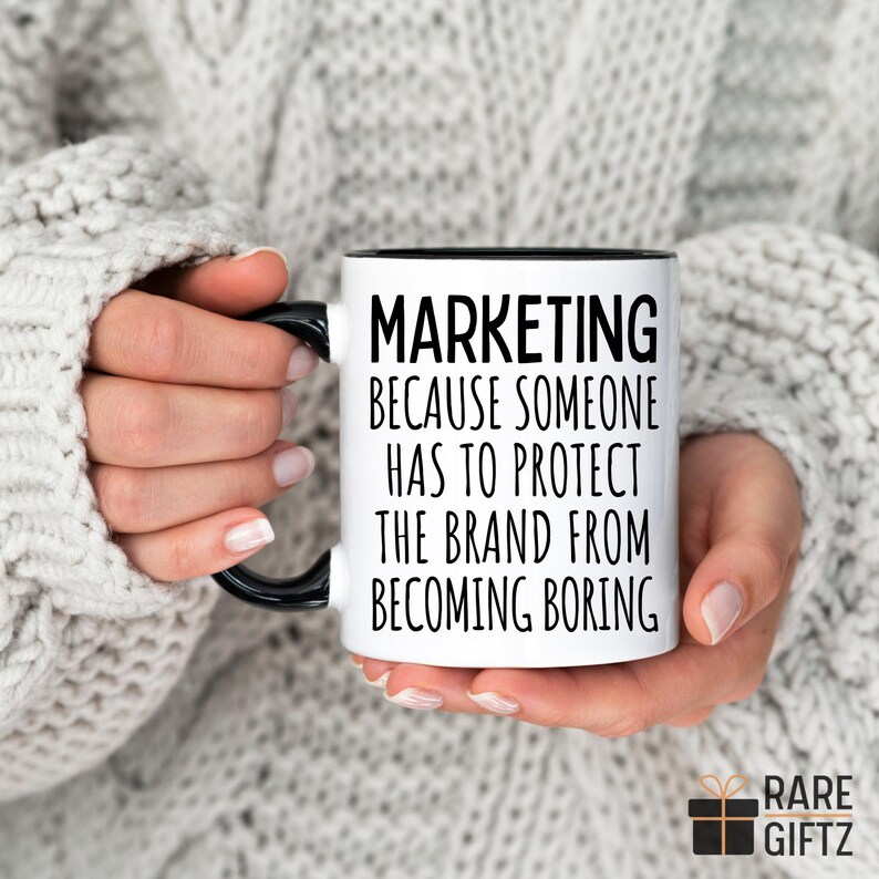 May include: White coffee mug with a black rim and handle. The mug has black text that reads "Marketing because someone has to protect the brand from becoming boring."