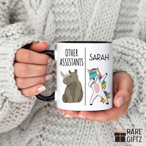 Peut inclure: Une tasse en c&eacute;ramique blanche avec un bord et une poign&eacute;e noirs. La tasse pr&eacute;sente un graphique d'un rhinoc&eacute;ros et d'une licorne avec le texte "Other Assistants" et "Sarah".